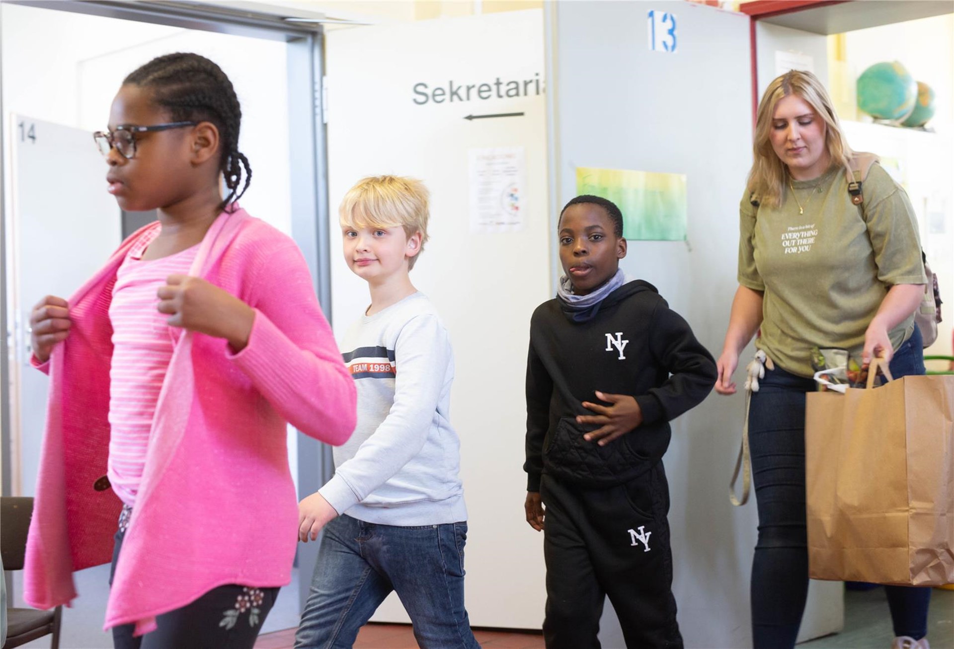 Vier Kinder gehen in einem Flur nebeneinander her. Links trägt ein Mädchen einen pinken Strickcardigan über einem gestreiften Shirt, daneben gehen ein Junge und ein Mann mit einer Papiertasche.