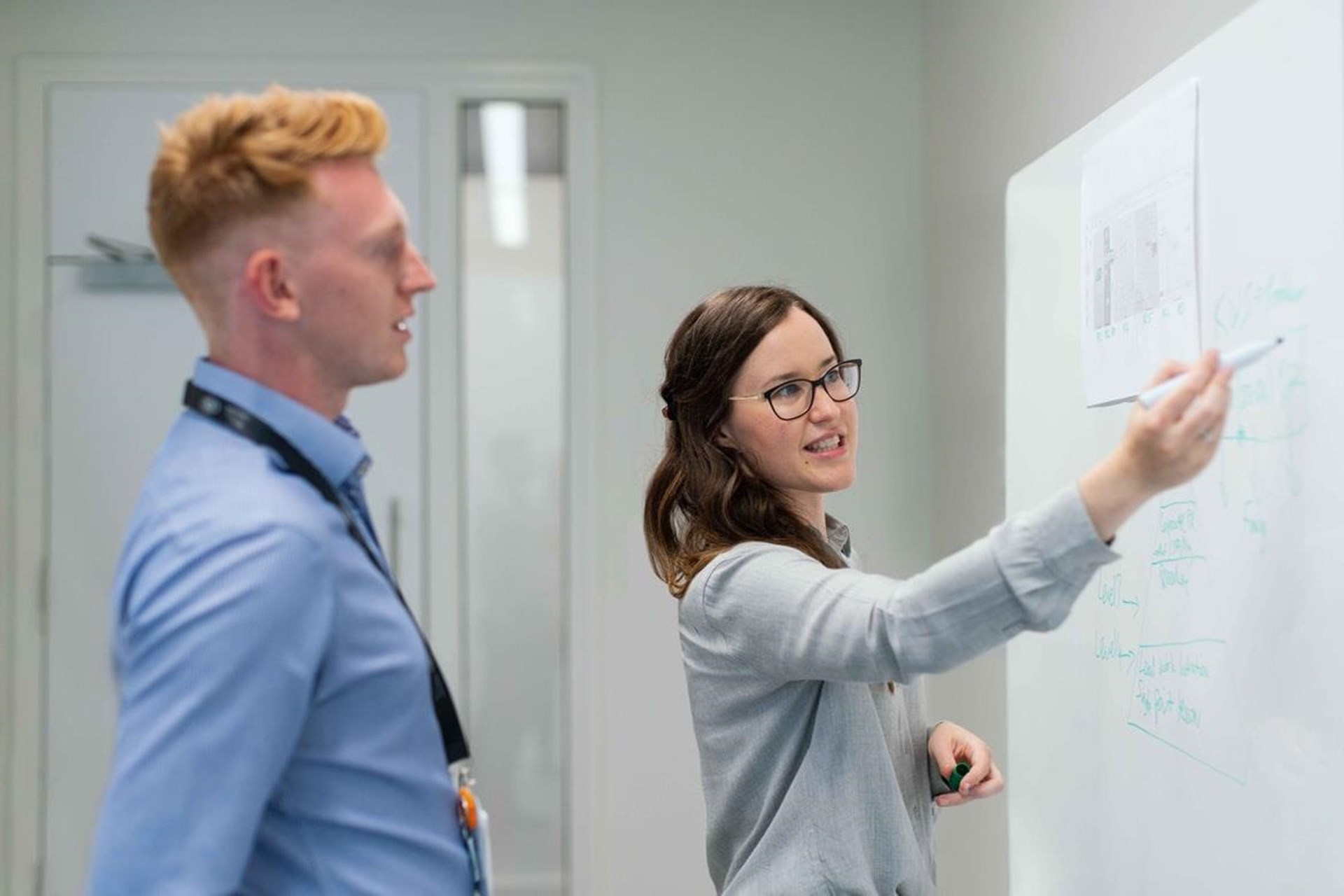 Eine Frau mit Brille erklärt an einem Whiteboard, während ein Mann in einem blauen Hemd aufmerksam zuhört.