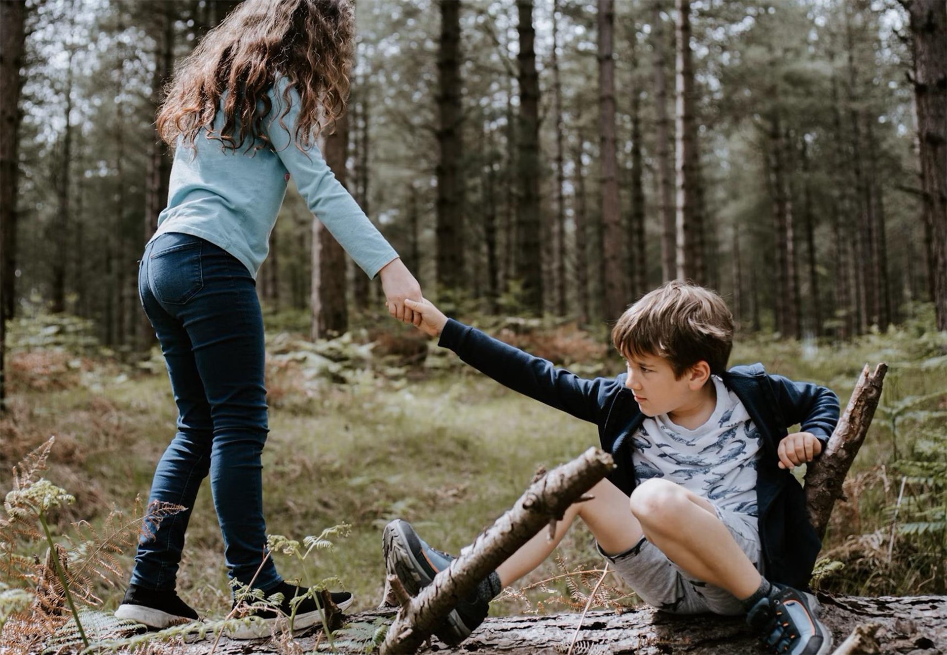 Ein Mädchen hilft einem Jungen, der auf einem Baumstamm im Wald sitzt, sich hochzuziehen.
