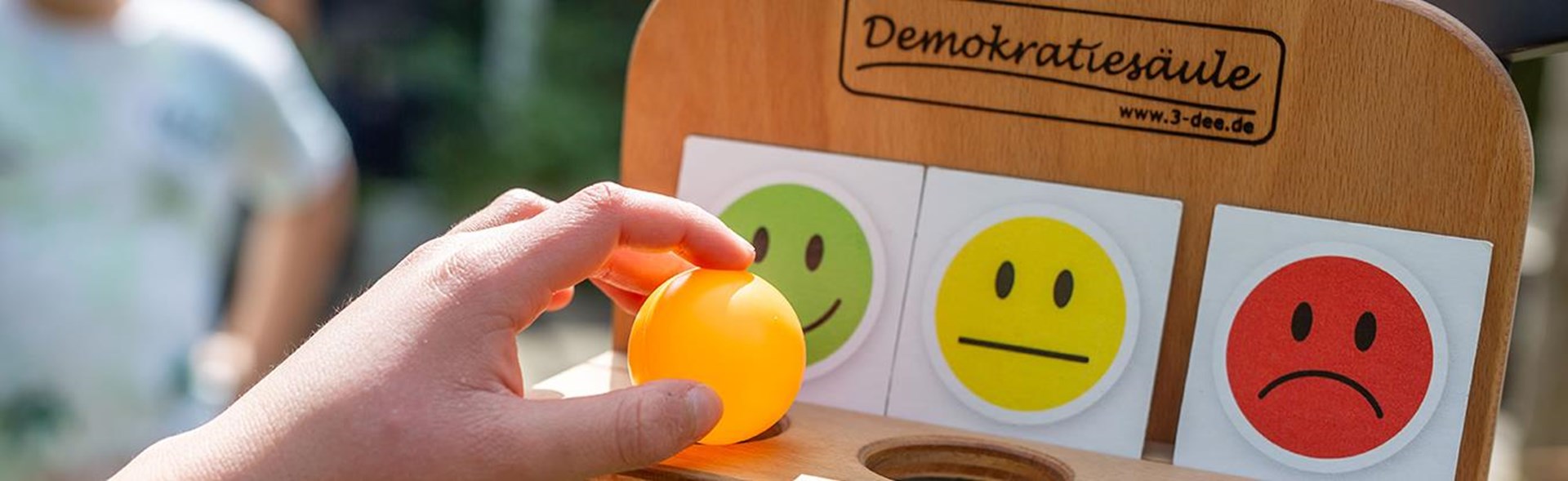 Eine Hand hält einen orangefarbenen Ball, während sie ihn über eine Holztafel mit zwei lächelnden Gesichtern positioniert.